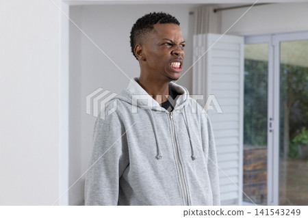 African american man standing at home next to sliding glass door pulling grimace in grey hoodie 141543249