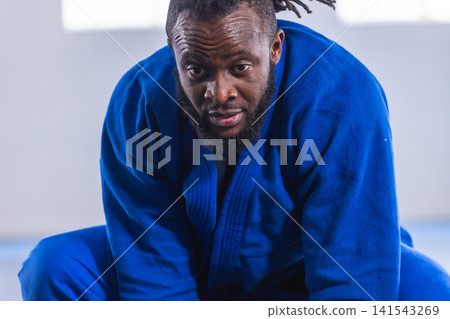 African american man kneeling on blue padded mat inside martial arts studio in blue gi African american man kneeling on blue padded mat inside martial arts studio in blue gi 141543269