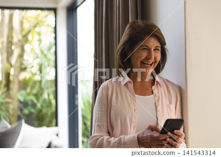 Senior woman leaning in living room by glass door, smiling and holding smartphone, copy space Senior woman leaning in living room by glass door, smiling and holding smartphone, copy space 141543314