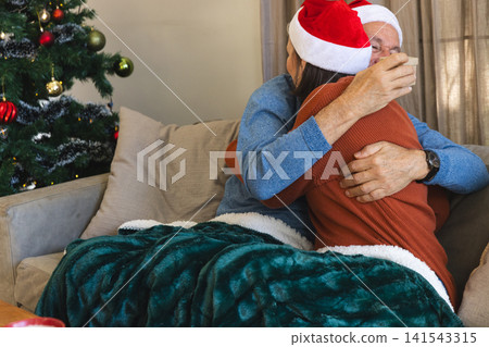 Senior father and daughter wearing Santa hats hugging on couch beside Xmas tree with red ornaments 141543315