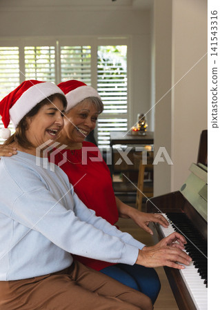 Senior female friends wearing Santa hats playing grand piano in living room with festive candle 141543316