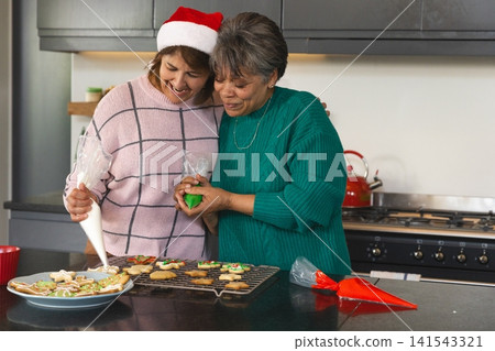 Diverse mother and daughter decorating holiday cookies on dark stone countertop with piping bags Diverse mother and daughter decorating holiday cookies on dark stone countertop with piping bags 141543321