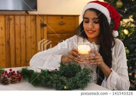 Asian woman sitting in living room making wreath holding lit candle wearing sweater and Santa hat Asian woman sitting in living room making wreath holding lit candle wearing sweater and Santa hat 141543328