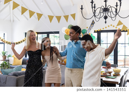 Diverse friends standing under gold pennant banners with balloons in living room Diverse friends standing under gold pennant banners with balloons in living room 141543351