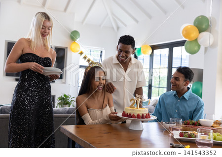 Diverse friends celebrating birthday party around wooden table with gold topper cake, balloons Diverse friends celebrating birthday party around wooden table with gold topper cake, balloons 141543352