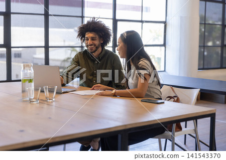 Diverse coworkers leaning forward and smiling while reviewing papers on laptop in office 141543370