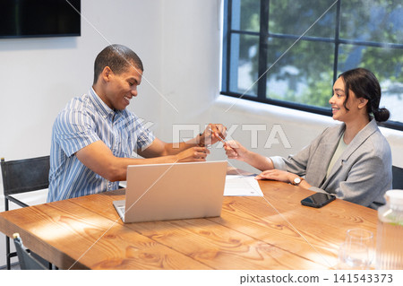 Diverse coworkers exchanging pen while sitting at conference table with laptop, coffee cup Diverse coworkers exchanging pen while sitting at conference table with laptop, coffee cup 141543373