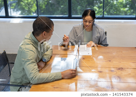 Diverse coworkers in business attire sitting at office table reviewing printed document, laptop Diverse coworkers in business attire sitting at office table reviewing printed document, laptop 141543374