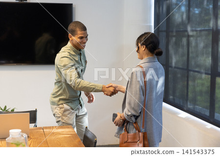 Diverse business associates shaking hands at conference room table, laptop, smartphone, bag Diverse business associates shaking hands at conference room table, laptop, smartphone, bag 141543375