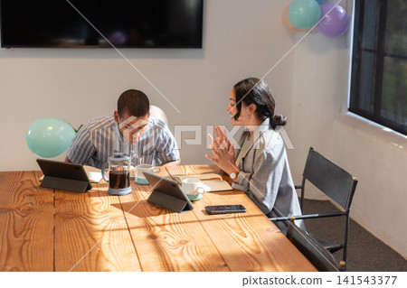 Diverse coworkers sitting at conference table using tablets with french press under balloons Diverse coworkers sitting at conference table using tablets with french press under balloons 141543377