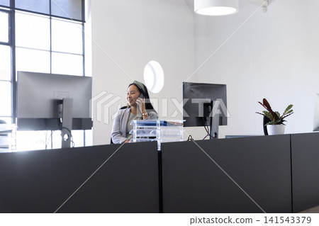 Asian woman wearing grey blazer sitting at desk partition using computer, phone, trays in office Asian woman wearing grey blazer sitting at desk partition using computer, phone, trays in office 141543379