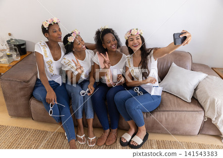 Diverse bridal party posing for selfie on living room sofa wearing sashes and holding smartphone Diverse bridal party posing for selfie on living room sofa wearing sashes and holding smartphone 141543383