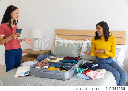 Diverse mom and teenage daughter packing gray suitcase on bed near bedside lamp Diverse mom and teenage daughter packing gray suitcase on bed near bedside lamp 141543386