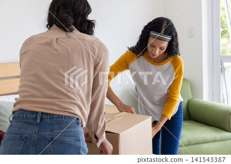 Diverse mom and teenage daughter leaning over cardboard box on bed using tape dispenser Diverse mom and teenage daughter leaning over cardboard box on bed using tape dispenser 141543387