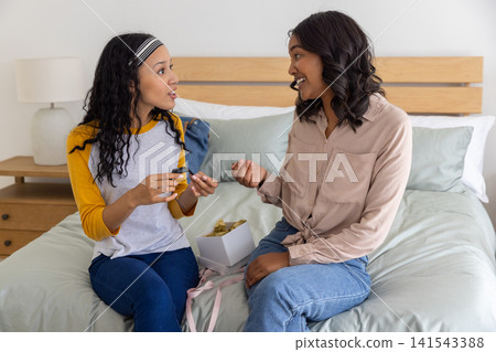 Diverse mom and teenage daughter sitting on bed in bedroom unpacking gift box and testing cosmetics 141543388