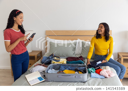 Diverse mom and teenage daughter sorting clothes and using smartphone in bedroom with suitcase Diverse mom and teenage daughter sorting clothes and using smartphone in bedroom with suitcase 141543389