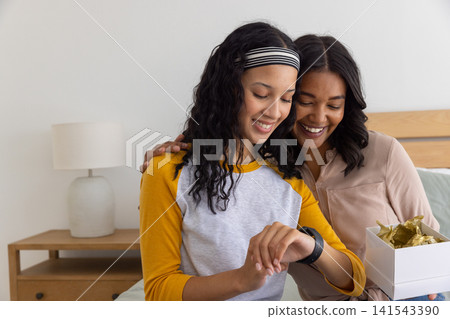 Diverse mom and teenage daughter sitting on bed examining smartwatch with gift box nearby Diverse mom and teenage daughter sitting on bed examining smartwatch with gift box nearby 141543390