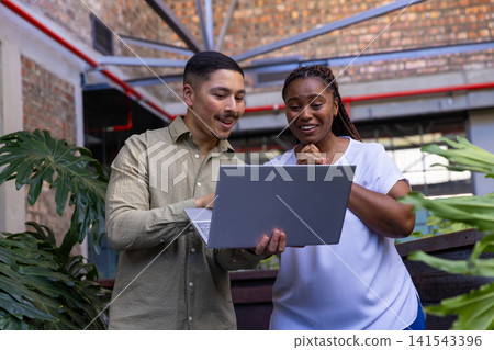 Diverse coworkers pointing at laptop in atrium with metal beams, railing and potted plants Diverse coworkers pointing at laptop in atrium with metal beams, railing and potted plants 141543396