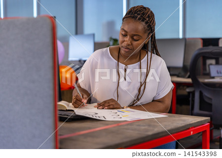 African american woman writing notes on colorful charts at office with balloons and monitors African american woman writing notes on colorful charts at office with balloons and monitors 141543398
