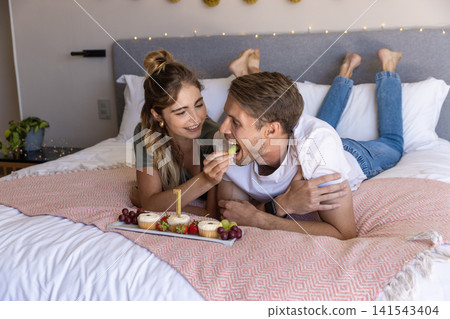 Couple lying side by side on zigzag blanket in bedroom feeding grapes alongside cupcakes Couple lying side by side on zigzag blanket in bedroom feeding grapes alongside cupcakes 141543404