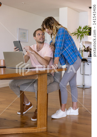 Couple collaborating on tablet while sitting at wooden dining table with laptop coffee mug papers Couple collaborating on tablet while sitting at wooden dining table with laptop coffee mug papers 141543406