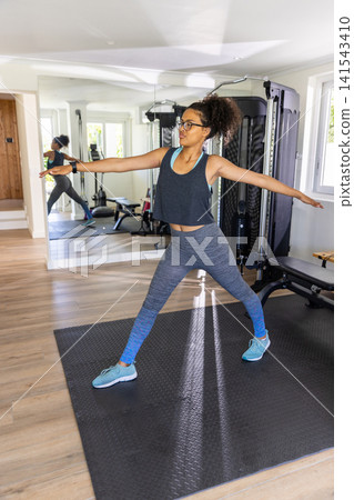 African American woman performing warrior II pose in sportswear on mat in home gym with mirror African American woman performing warrior II pose in sportswear on mat in home gym with mirror 141543410
