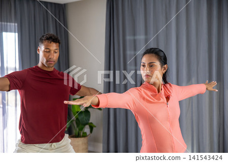 Diverse workout partners practicing warrior ii pose in home living room by plant 141543424