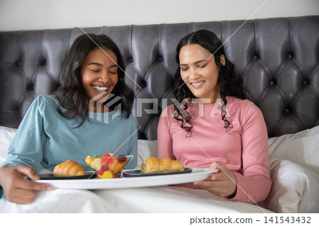 Diverse female couple sharing breakfast tray on bed with tufted headboard, croissants, fruit Diverse female couple sharing breakfast tray on bed with tufted headboard, croissants, fruit 141543432