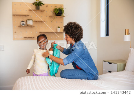 African american mother kneeling on bed in bedroom, helping daughter adjust turquoise backpack African american mother kneeling on bed in bedroom, helping daughter adjust turquoise backpack 141543442
