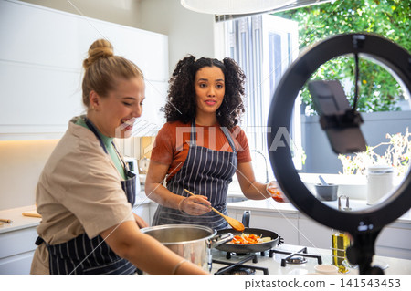 Diverse female friends stirring, cooking chopped vegetables on kitchen island with ring light Diverse female friends stirring, cooking chopped vegetables on kitchen island with ring light 141543453