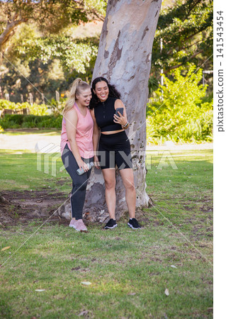 Diverse female friends leaning against tree at park taking selfie with phone, water bottle 141543454