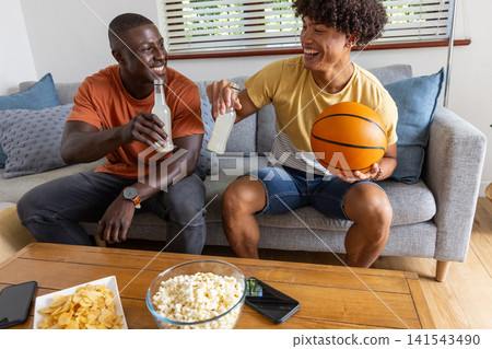 Diverse male friends clinking drinks on couch near coffee table with basketball and snacks Diverse male friends clinking drinks on couch near coffee table with basketball and snacks 141543490