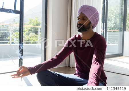 Meditating man sitting on yoga mat next to sliding glass door, inhaling incense smok Meditating man sitting on yoga mat next to sliding glass door, inhaling incense smok 141543519
