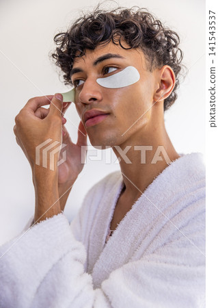 Man applying hydrogel under-eye patch while wearing plush white bathrobe at bathroom Man applying hydrogel under-eye patch while wearing plush white bathrobe at bathroom 141543537