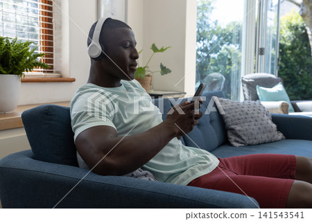 African american man tapping smartphone in living room by sliding door, wearing headphones African american man tapping smartphone in living room by sliding door, wearing headphones 141543541