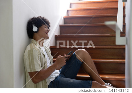 African american male on steps wearing headphones smiling at smartphone, copy space 141543542