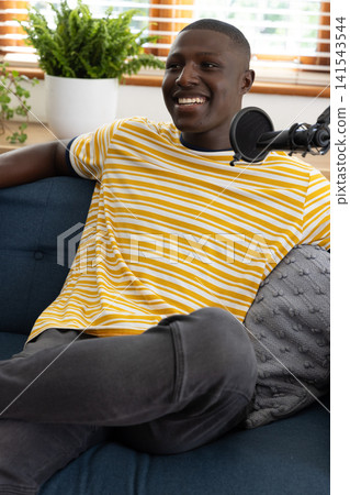 African american man speaking into microphone, sitting on couch by fern in living room African american man speaking into microphone, sitting on couch by fern in living room 141543544
