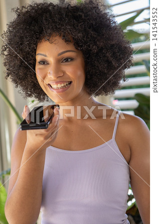 Adult african american woman holding smartphone at mouth and speaking at home with plants by window Adult african american woman holding smartphone at mouth and speaking at home with plants by window 141543552