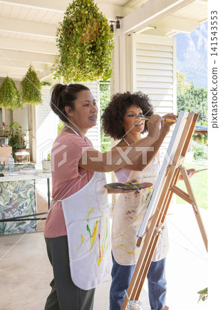 Asian instructor guiding her student painting on wooden easel at covered porch near plant Asian instructor guiding her student painting on wooden easel at covered porch near plant 141543553