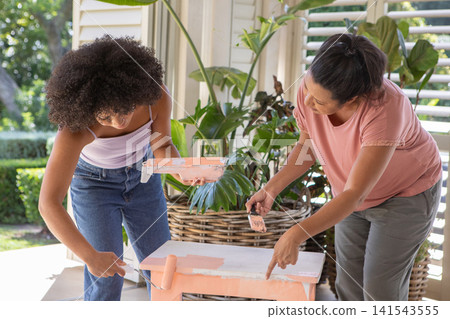 Diverse mother and daughter painting wooden table on drop cloth on porch with paint with roller Diverse mother and daughter painting wooden table on drop cloth on porch with paint with roller 141543555