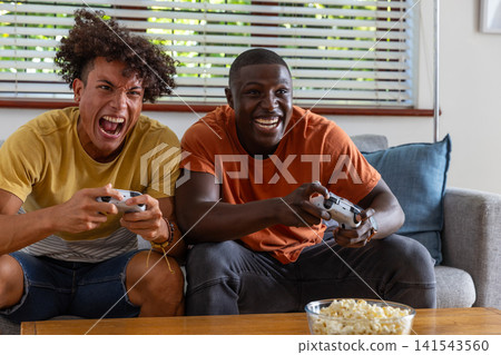 Diverse male friends leaning forward on gray sofa in living room holding controllers, popcorn Diverse male friends leaning forward on gray sofa in living room holding controllers, popcorn 141543560