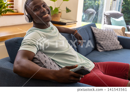 African american man relaxing on couch at home wearing headphones holding smartphone 141543561