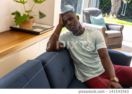 African american man sitting on couch leaning head on hand inside living room African american man sitting on couch leaning head on hand inside living room 141543563