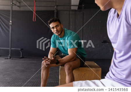 Male gym partners wearing workout tees chatting on plyometric box at gym with red resistance band Male gym partners wearing workout tees chatting on plyometric box at gym with red resistance band 141543568