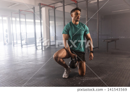 Kneeling man wearing teal shirt gripping black kettlebell on gym floor near squat rack, copy space Kneeling man wearing teal shirt gripping black kettlebell on gym floor near squat rack, copy space 141543569