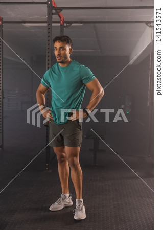 Man standing with hands on hips in gym beside power rack wearing teal shirt and smartwatch Man standing with hands on hips in gym beside power rack wearing teal shirt and smartwatch 141543571