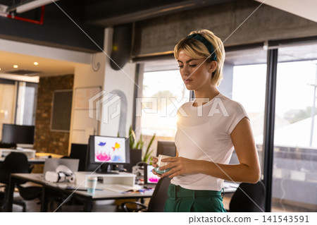 Woman standing in office workspace with ceramic mug reviewing computer monitors, copy space Woman standing in office workspace with ceramic mug reviewing computer monitors, copy space 141543591