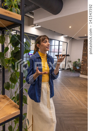 Mid adult woman holding smartphone and gesturing near metal shelving unit with potted plants Mid adult woman holding smartphone and gesturing near metal shelving unit with potted plants 141543592