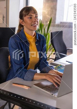 Mid adult woman wearing denim shirt typing on laptop at office desk with metallic pen 141543593