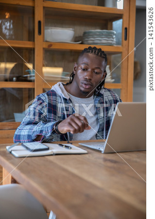 African american man leaning forward, reaching toward notebook at home dining table with laptop African american man leaning forward, reaching toward notebook at home dining table with laptop 141543626